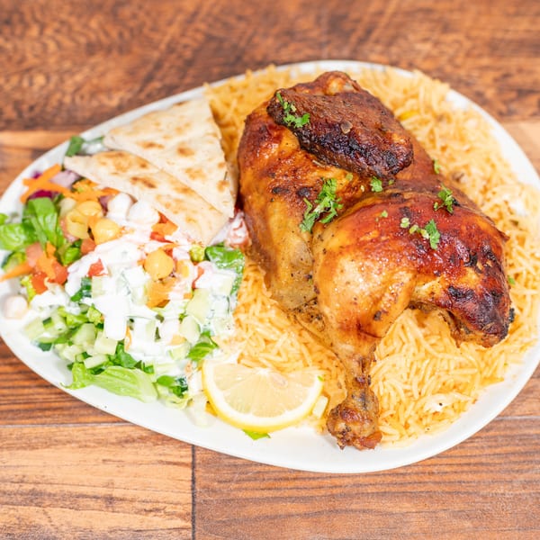Grilled chicken leg quarter with rice, salad, pita bread, and a lemon wedge on a white plate.