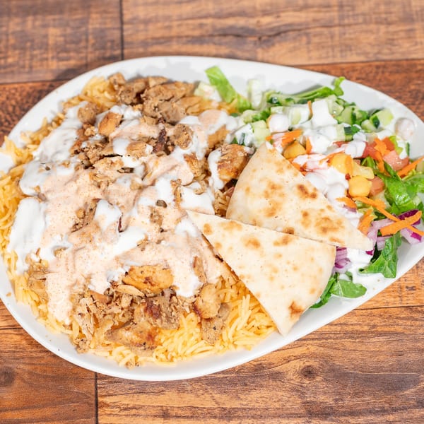 Chicken over rice with white sauce, hot sauce, salad, and pita bread on a white plate.