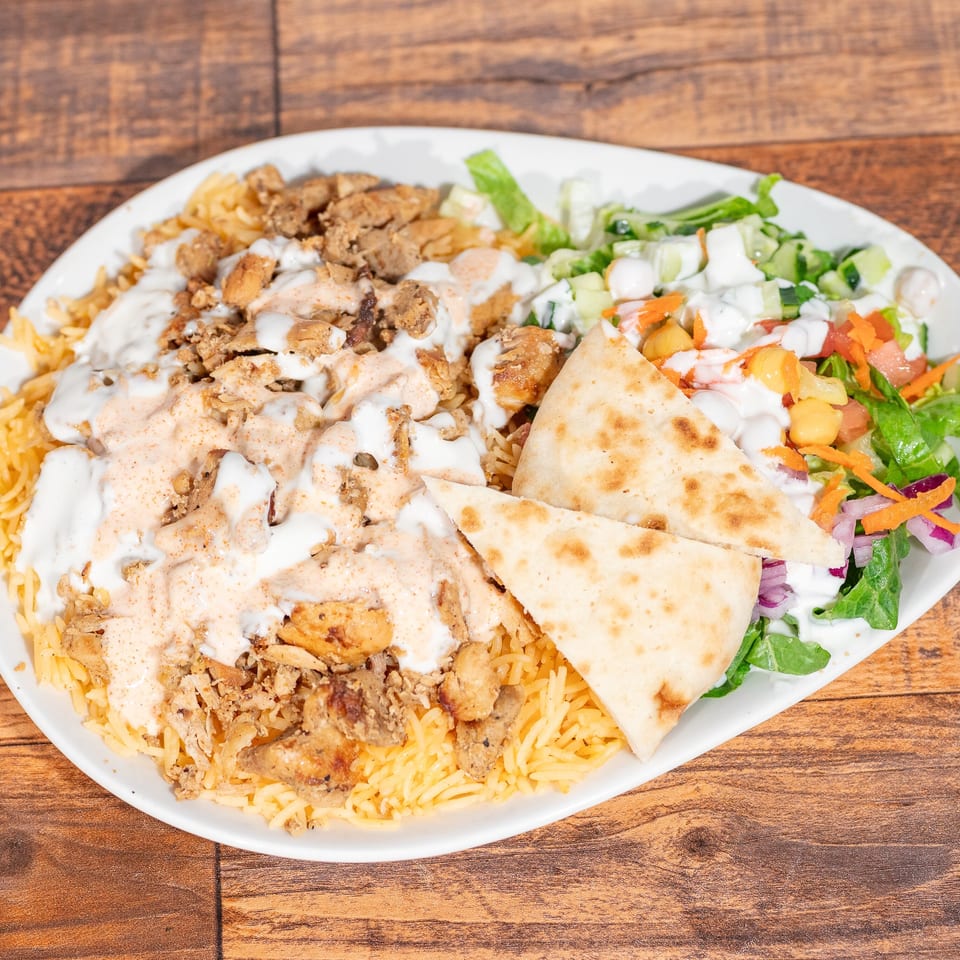 Chicken over rice with white sauce, hot sauce, salad, and pita bread on a white plate.