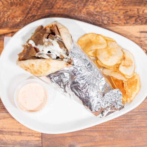 Gyro sandwich wrapped in foil with a side of potato chips and dipping sauce on a white plate.