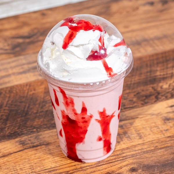 Strawberry milkshake with whipped cream and strawberry syrup in a clear plastic cup on a wooden table.
