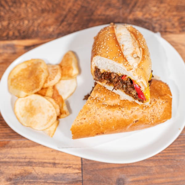 A sliced steak and pepper sandwich with chips on a white plate, on a wooden table.